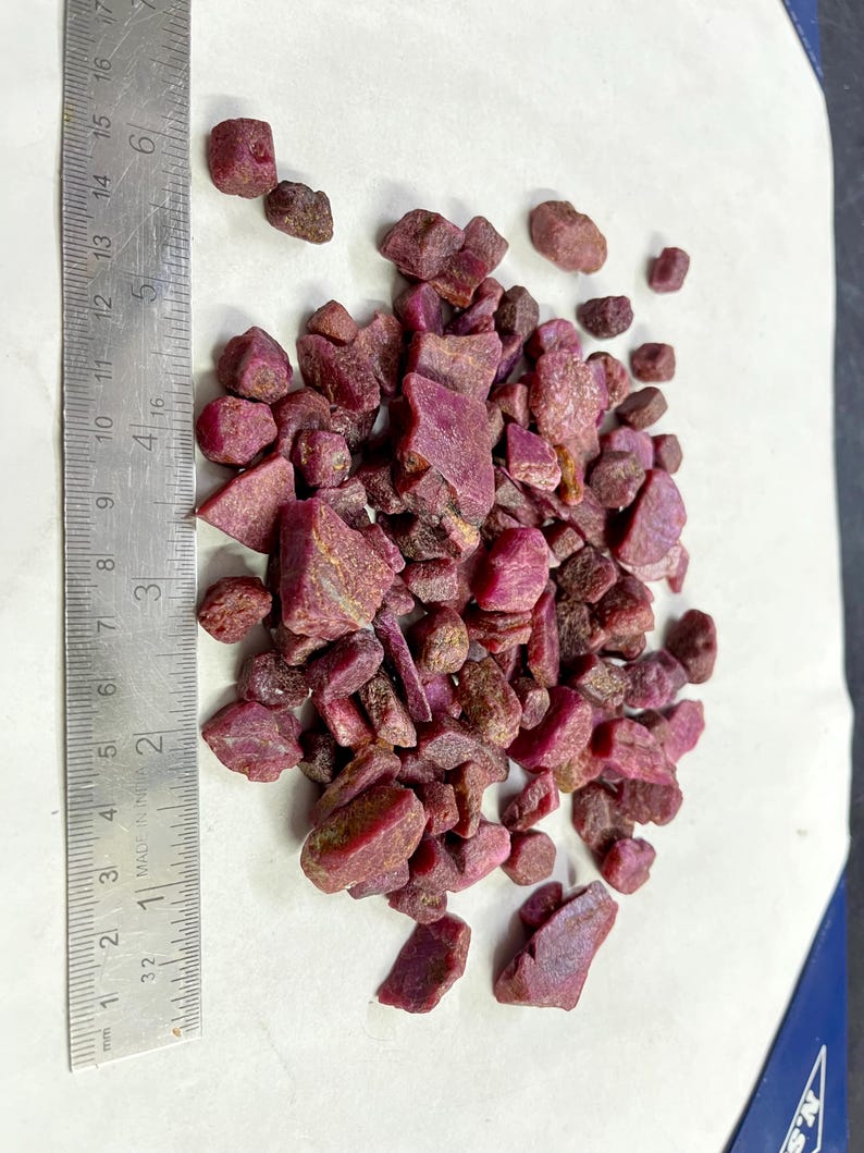 Op de afbeelding: Een verzameling ruwe, ongeslepen robijn edelstenen in verschillende maten en vormen. De dieprode tot paarse stenen liggen verspreid op een wit oppervlak naast een metalen liniaal.