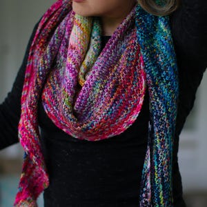 May include: A hand-knitted triangular shawl featuring a vibrant, multi-coloured variegated yarn pattern. The knit fabric displays a dense, textured stitch with a gradient transition from bright pinks and oranges to deep blues and teals.