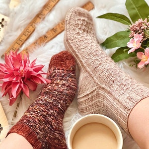 May include: Two pairs of hand-knitted socks in different colors and patterns. One pair is a light brown with a textured stitch, the other is a multi-colored yarn with a more intricate pattern. The socks are on a person's feet, with a cup of coffee in the foreground.