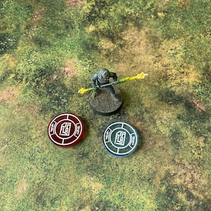 May include: A miniature figure of a soldier holding a yellow staff, standing on a black base. Two circular tokens with a white logo and red and blue borders are on a green and brown surface.