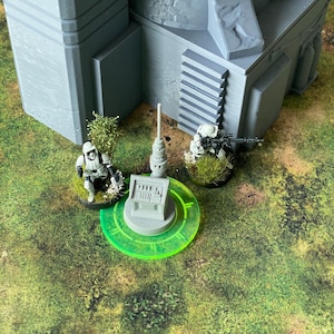 Può includere: Un modello grigio in miniatura di un edificio con due figure di Stormtrooper e una figura di cecchino su una superficie verde e marrone. Una base circolare verde con una struttura grigia è in primo piano. La scena è ambientata su un terreno erboso.