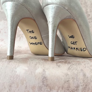 May include: A pair of silver high-heeled shoes with the phrase "The One Where" written on one shoe and "We Get Married" written on the other shoe.