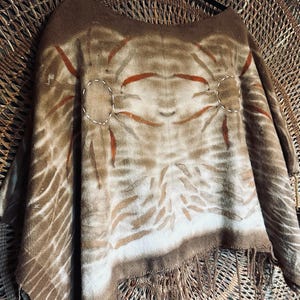 May include: A brown poncho featuring a textured, fuzzy surface with a tie-dye pattern in shades of tan, cream, and rust orange. The garment includes two circular stitched accents on the upper chest and a fringed hem along the bottom edge.
