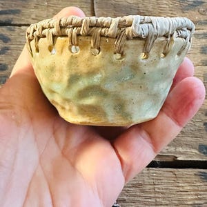 May include: A small, handmade ceramic bowl with a woven rim. The bowl has a light green and tan colour scheme with a textured surface. The rim is made of woven natural fibres, adding a rustic touch to the piece.