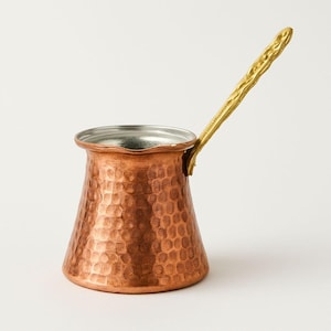 Op de afbeelding: Een koperkleurige Turkse koffiepot met een gehamerde textuur en een goudkleurige handgreep. De pot heeft een brede basis die taps toeloopt naar een smallere opening. De handgreep is lang en omhoog gericht.