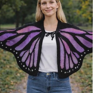 Op de afbeelding: Een zwarte en paarse gebreide sjaal in de vorm van vlindervleugels met witte sterren. De sjaal heeft een zwarte striksluiting en wordt gedragen over een wit shirt en een blauwe spijkerbroek. Een uniek modeaccessoire.