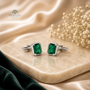 May include: A pair of silver cufflinks with emerald-cut, vibrant green gemstones. The cufflinks are displayed on a light-colored, marble-like surface, with a soft, neutral-toned background. The text "PEACOCK JEWEL COLLECTION 24" is visible in the upper left corner.