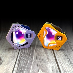 Puede incluir: Dos contadores de juego hexagonales, uno de color morado claro y otro naranja, cada uno con un inserto central iridiscente multicolor. El contador morado muestra el número 0, mientras que el contador naranja muestra el número 9 en blanco.