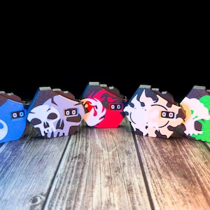 Puede incluir: Una fila de cinco máscaras decorativas con diseños únicos. Las máscaras presentan una gota de agua azul, una calavera, una llama roja, un patrón blanco y negro y una nube verde. Cada máscara tiene un marco marrón oscuro y dos pequeños agujeros cuadrados para los ojos.