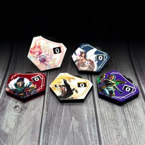 Könnte beinhalten: Fünf sechseckige Spielmarken mit Charakterkunst und der Zahl "0". Die Marken haben einen schwarzen Rand und liegen auf einer Holzoberfläche. Die Kunst zeigt Charaktere in verschiedenen Farben.