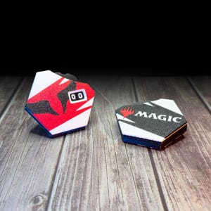 Puede incluir: Dos contadores de juego hexagonales. Uno es blanco con detalles en rojo y negro y una pantalla digital que muestra "00". El otro es blanco y negro con la palabra "MAGIC". Ambos tienen bordes azules y negros. Los contadores están sobre una superficie de madera.