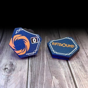 May include: Two blue hexagonal game tokens with white trim. One token has an orange swirl design and the number 0. The other token has the text "RIFTBOUND" in orange. Both tokens are on a wooden surface.