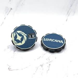 Puede incluir: Dos fichas de juego azul marino y negro. Una ficha muestra un emblema blanco y el número 20. La otra ficha tiene la palabra "LORCANA" en blanco. Ambas fichas tienen un borde negro estriado.