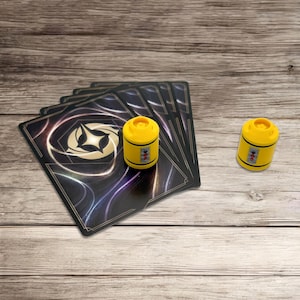 Puede incluir: Un mazo de cinco cartas de juego rectangulares con fondo negro, líneas blancas en espiral y un emblema de estrella dorada central, acompañado por dos pequeñas fichas cilíndricas de plástico amarillo con un detalle gráfico vertical en rojo y plata.