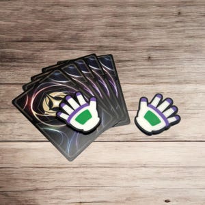 May include: A collection of playing cards with a purple and gold design, accompanied by two hand-shaped charms. The charms are white with purple outlines and a green center, resting on a wooden surface.