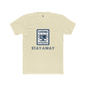 Könnte beinhalten: Cremefarbenes T-Shirt mit marineblauem Aufdruck, der "CODING IN PROGRESS" über dem Wort "STAY AWAY" zeigt. Die Grafik zeigt ein Computermonitor-Symbol. Das Shirt hat einen Rundhalsausschnitt und kurze Ärmel.