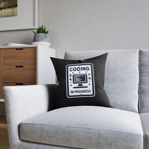 May include: Black square cushion featuring a white and navy blue graphic that reads "CODING IN PROGRESS". The cushion is placed on a grey sofa. The graphic includes a computer monitor icon and other symbols.
