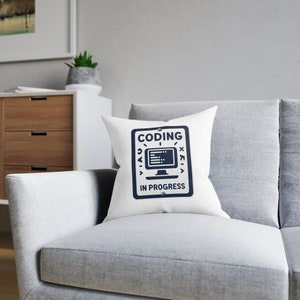 May include: White square cushion featuring a navy blue graphic with the text "CODING IN PROGRESS". The design includes a computer monitor icon and other symbols. The cushion is placed on a grey sofa.