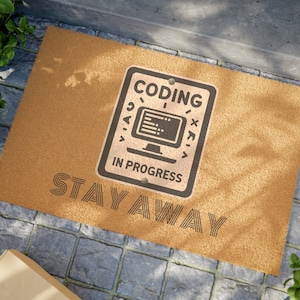 May include: A rectangular doormat made of tan coir fiber. The mat features a black and white sign that reads "CODING IN PROGRESS" with a computer graphic. Below the sign, the words "STAY AWAY" are printed in black.