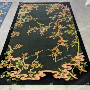 May include: A rectangular black rug with an embroidered floral design. The rug features branches with green leaves, orange and red flowers, and a light brown outline. The design is set against a dark green background.