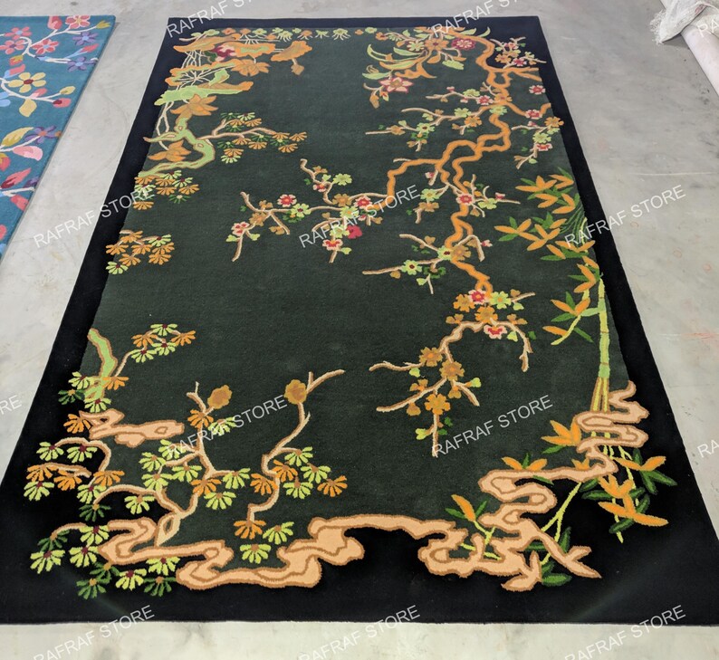 May include: A rectangular black rug with an embroidered floral design. The rug features branches with green leaves, orange and red flowers, and a light brown outline. The design is set against a dark green background.