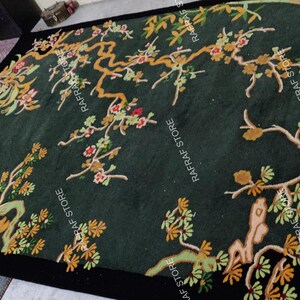 May include: A dark green rug with a black border, featuring an embroidered design of flowering branches in shades of gold, green, and red. The rug has a rectangular shape and is placed on a light-coloured surface.