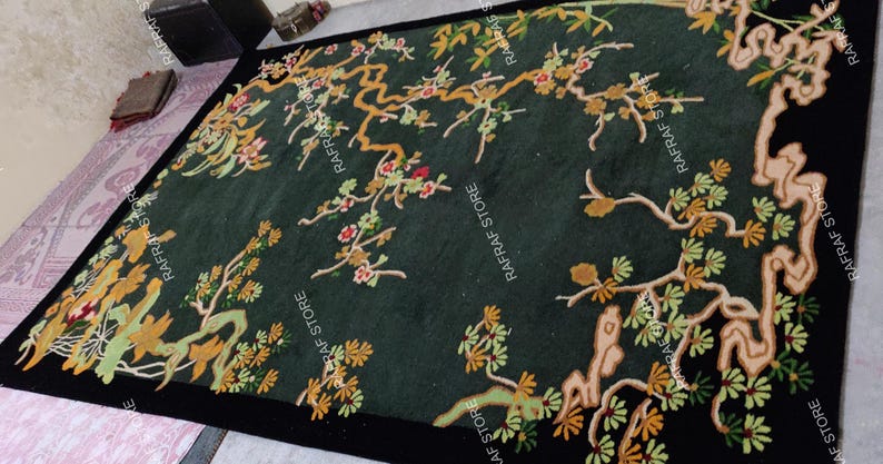 May include: A dark green rug with a black border, featuring an embroidered design of flowering branches in shades of gold, green, and red. The rug has a rectangular shape and is placed on a light-coloured surface.