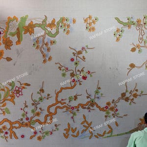 May include: An off-white textile with an embroidered floral pattern in orange, green, and red. The design includes branches, flowers, and leaves. The words "RAFRAF STORE" are printed on the textile.