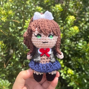 Peut inclure: Une poupée crochetée avec des cheveux bruns ornés d'un nœud blanc, des yeux verts et une petite bouche brodée. La poupée porte un blazer gris sur un haut marron, un nœud papillon rouge, une jupe plissée bleu foncé et des chaussures noires.