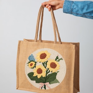 Peut inclure: Un sac fourre-tout en jute brun avec un motif circulaire brodé de tournesols et de feuilles. Les tournesols sont jaunes avec des centres bruns et les feuilles sont vertes. Un ruban rose est noué autour des tiges des tournesols.