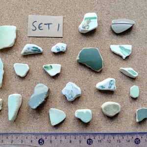 May include: A collection of twenty-one assorted sea glass and ceramic shards in various shades of green and white, some featuring painted patterns. A small rectangular card labelled SET 1 is included, and a ruler shows a scale of 18 centimetres.