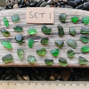 May include: A selection of green sea glass pieces displayed on a weathered wooden plank. The sea glass varies in shape and shade, from light to dark green. A small card with "SET 1" is visible. The arrangement is on a bed of pebbles.