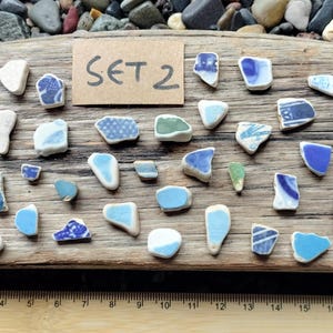 May include: A display of sea pottery fragments, predominantly blue and white, arranged on driftwood. The pieces vary in shape and size. A small card reads "SET 2". A wooden ruler, marked in centimetres, is at the bottom of the image.