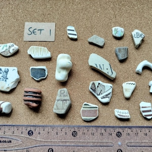 May include: A collection of twenty-one assorted ceramic shards and pottery fragments featuring various patterns, colours, and textures, including one piece with the text 'SET 1', arranged above a clear plastic ruler measuring up to 21 centimetres.