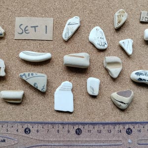 May include: A collection of twenty-one assorted white and off-white ceramic sea pottery shards, some featuring black printed text or patterns, arranged near a rectangular card labelled SET 1 and a clear plastic ruler measuring up to 20 cm.