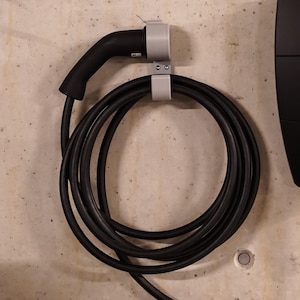 Op de afbeelding: Een zwarte elektrische voertuiglader aan een betonnen muur. De lader heeft een groene lichtcirkel met een witte 'Z' in het midden. Een zwarte laadkabel is opgerold en wordt vastgehouden door een grijze beugel.