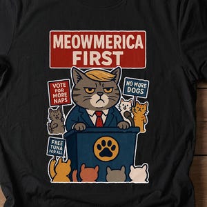 May include: Black t-shirt with a cartoon cat wearing a suit and tie. The cat is standing behind a podium with the text "MEOWMERICA FIRST" on it. There are other cats around the podium with signs that say "VOTE FOR MORE NAPS", "NO MORE DOGS", and "FREE TUNA FOR ALL".