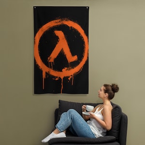 May include: Black wall flag with an orange lambda symbol in a circle, spray-painted effect. The rectangular flag hangs vertically. The design has a grunge aesthetic, with drips of orange paint. The flag is suitable for a bedroom or games room.