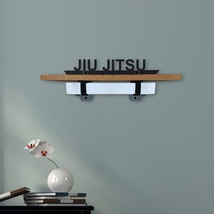 Op de afbeelding: Een houten plank met de zwarte tekst "JIU JITSU". Daaronder een witte band, vastgehouden door twee zwarte beugels. De plank is bevestigd aan een lichtblauwe muur.
