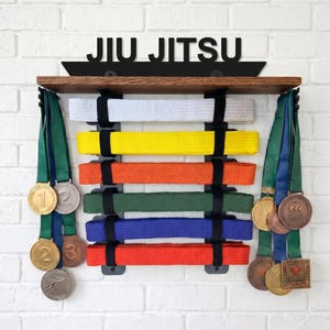 Puede incluir: Un estante de madera para cinturones y medallas de Jiu Jitsu, montado en una pared de ladrillos blancos. El estante exhibe cinturones blancos, amarillos, naranjas, verdes, azules y rojos. Las medallas cuelgan a ambos lados.
