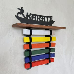 May include: A wooden shelf with a black metal "KARATE" sign and a martial artist silhouette. Below, six coloured karate belts are displayed: white, yellow, orange, green, blue, and red. The belts are mounted on the wall.