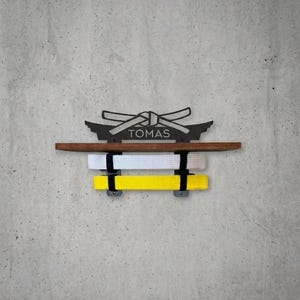 Pode incluir: Um expositor de parede para faixas de artes marciais com uma prateleira de madeira castanho-escura e um suporte de metal preto com o nome TOMAS gravado sob um gráfico de nó de faixa. O suporte segura uma faixa branca e uma amarela.