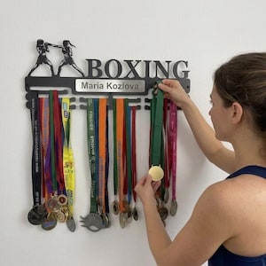 May include: Black metal boxing medal display with the word "BOXING" and a silhouette of two boxers. The display holds multiple colourful medals with ribbons. A person is placing a medal on the display.
