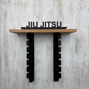 Op de afbeelding: Een houten plank met de zwarte tekst "JIU JITSU". De plank wordt ondersteund door twee zwarte beugels met rechthoekige uitsparingen. De achtergrond is een lichtgekleurde, getextureerde muur. Voor vechtsporten.