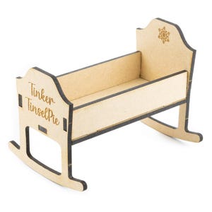 May include: A small, unfinished wooden rocking cradle with the text "Tinker TinselPie" on the side. The cradle has a snowflake design on the headboard. The cradle is made of light-colored wood with dark edges.