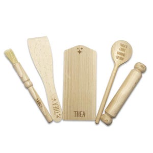 Può includere: Un set di utensili da cucina in legno per la cottura, tra cui un mattarello, una spatola, un pennello da pasticceria, un cucchiaio di legno e un tagliere. Gli utensili sono incisi con il nome "Thea's First Baking Spoon".
