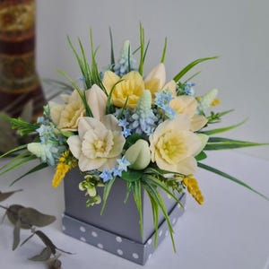 Peut inclure: Un bouquet de jonquilles jaunes et blanches, de myosotis bleus et de muscaris bleus disposés dans une boîte grise à pois blancs.