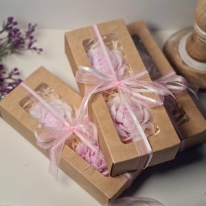 Può includere: Tre scatole regalo con finestre trasparenti, ciascuna contenente un sapone a forma di fiore rosa. Le scatole sono legate con un nastro rosa e sono impilate l'una sull'altra.