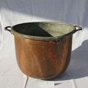 Puede incluir: Una olla grande de cobre con dos asas. La olla es vieja y tiene una pátina en la superficie. La olla está sobre una superficie blanca.