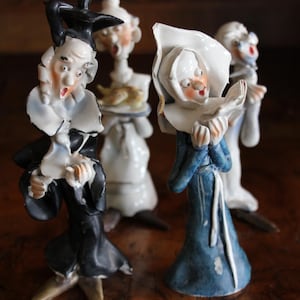 May include: A collection of vintage ceramic figurines, each depicting a character with exaggerated features. The figures are dressed in various outfits, including a blue dress and a black coat. The figurines have a glossy finish and are set against a dark wooden surface.